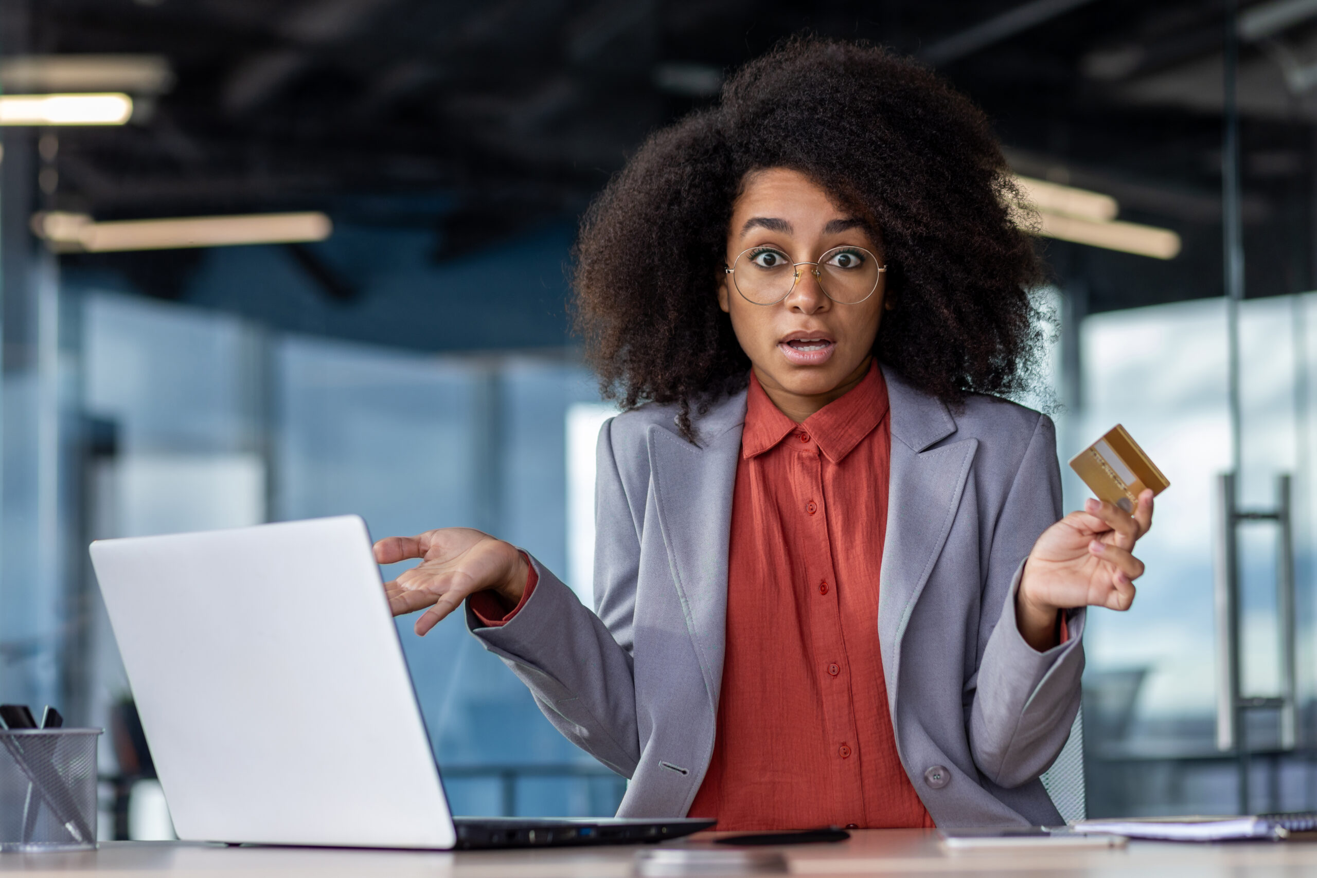 Shocked african american woman spreading hands with credit card while sitting by desk with laptop. Puzzled office worker facing struggles with online transaction due to weak connection in cabinet.